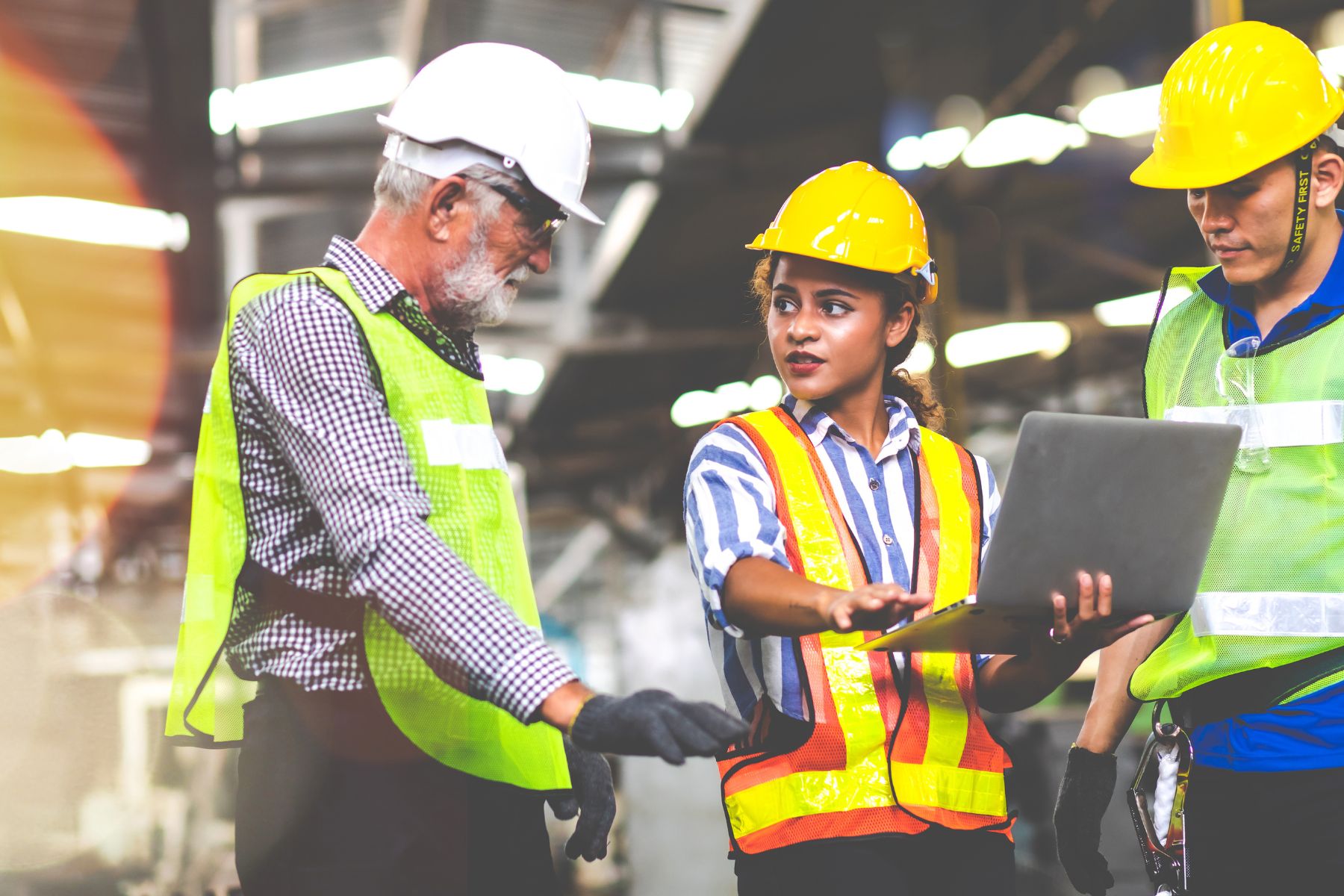 Three industrial workers wearing hard hats and safety vests review information on a laptop inside a manufacturing facility.